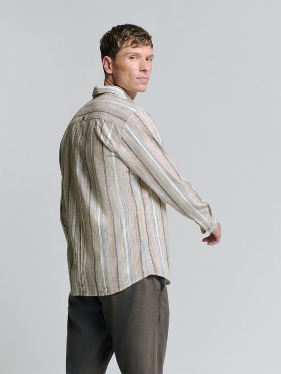 Model draagt een beige linnen mix blouse met verticale strepen en een witte tanktop. De blouse is half opengemaakt en de mouwen zijn opgerold.