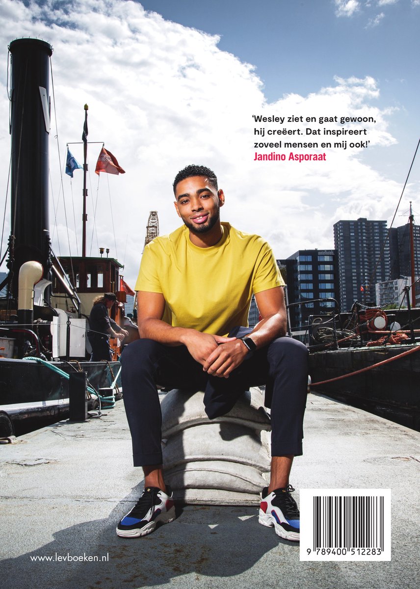 Jongeman met donker haar en baard, in een geel T-shirt en donkere broek, zit op een zak bij een boot.