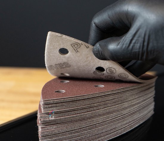 Hand met zwarte handschoen pakt bruin delta schuurpapier uit een stapel.