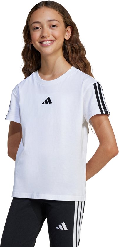 Jeune fille portant un t-shirt blanc avec logo adidas noir et trois bandes sur les manches, et un pantalon noir à bandes blanches.