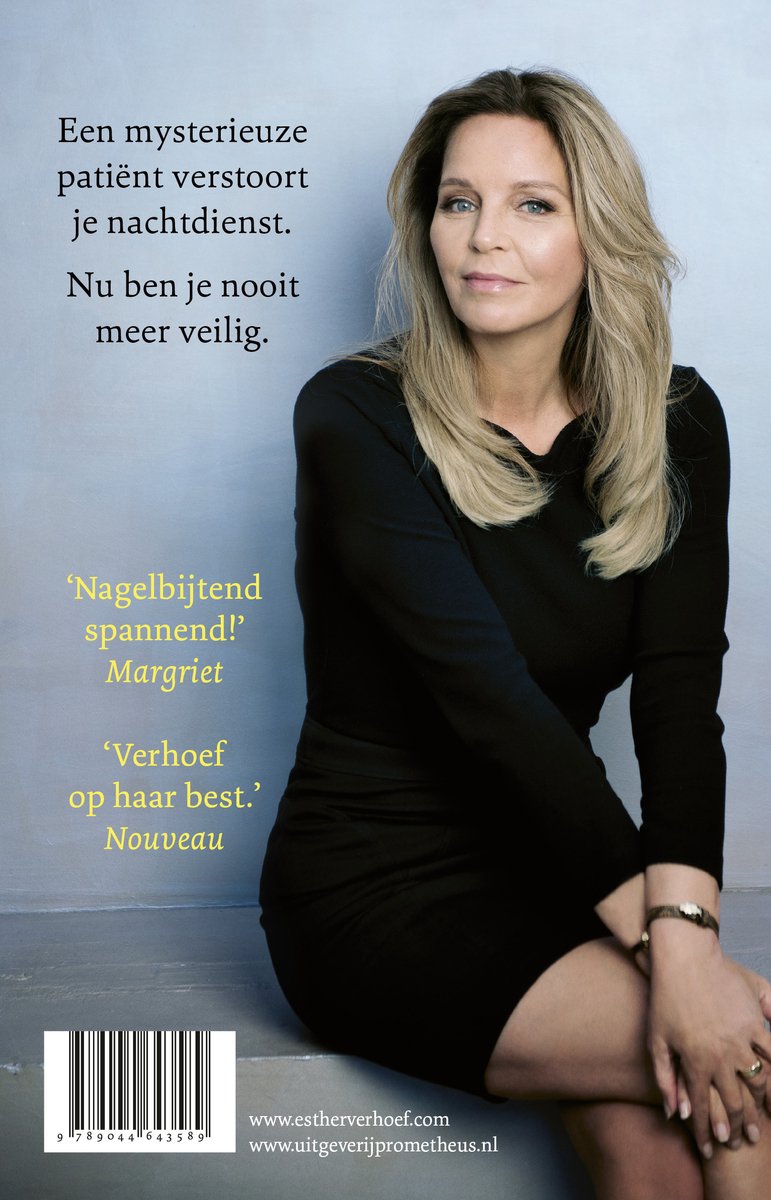 Een vrouw met blond haar, gekleed in een zwarte jurk, zit op een grijze achtergrond. Ze draagt een horloge.