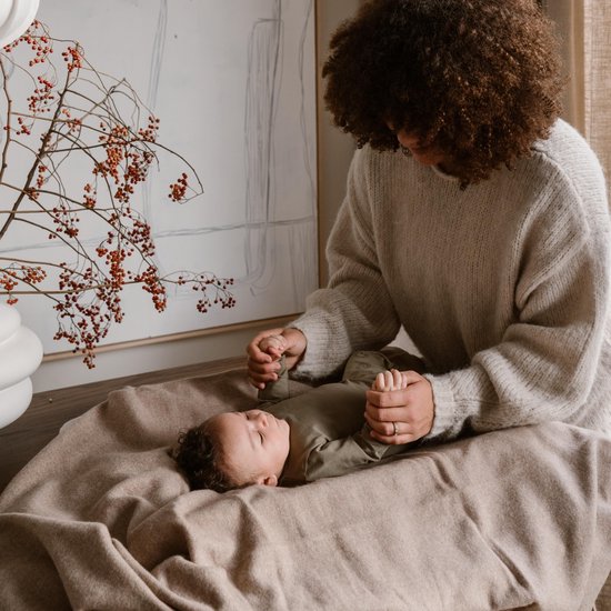 Een moeder met een lichte huid met krullend bruin haar in een beige trui buigt zich over haar baby die op een beige deken ligt.