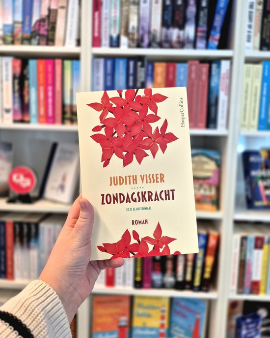 Hand houdt boek met lichtgele kaft en rode Ixora bloemen vast. Achtergrond boekenplank.