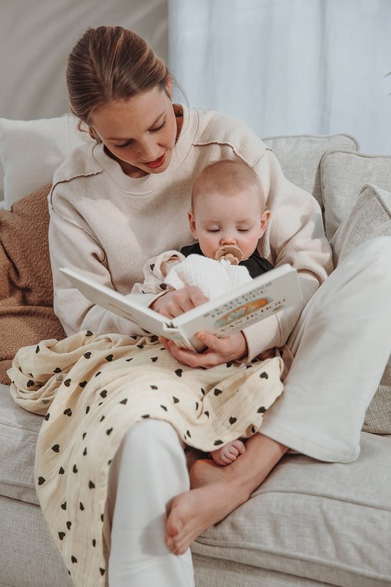 Vrouw leest boek aan baby die op haar schoot ligt, bedekt met een swaddle met hartjesprint.