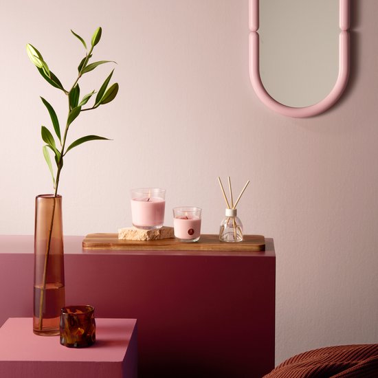 Gedecoreerd interieur met een glazen geurverspreider, twee roze kaarsen en een vaas met takken op een roze en bordeauxrode ondergrond.