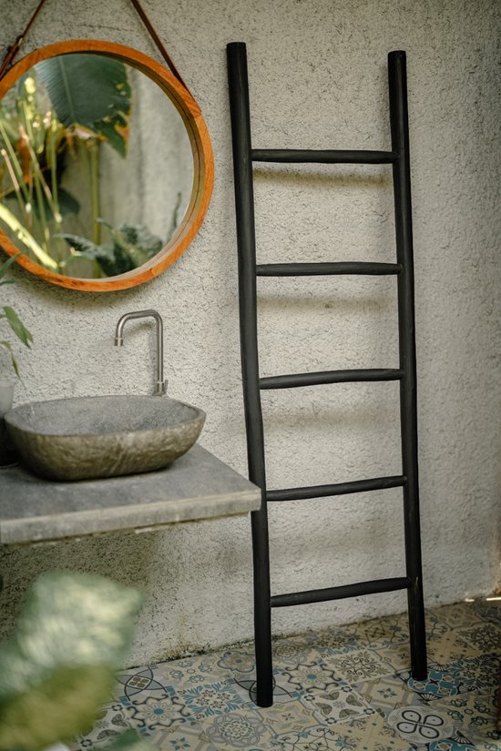 Zwarte houten decoratieve ladder staat naast een betonnen muur, naast een wastafel en ronde spiegel.