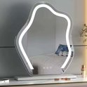 Golvende spiegel met ledverlichting op een witte basis, geplaatst op een kaptafel met make-up en een bed op de achtergrond.