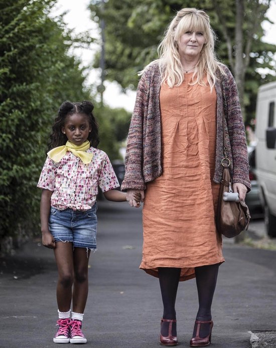 Een vrouw met blond haar, een oranje jurk en een gebreide vest, houdt de hand vast van een jong meisje met zwart haar in staartjes, een blouse en een spijkerbroek.