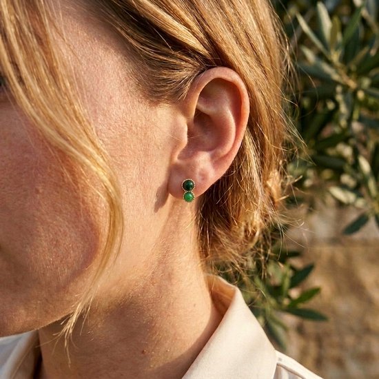 Model draagt gouden oorknopjes met twee ronde groene stenen in elk oor.