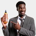 Glimlachende man in een pak wijst naar een fles Reuzel Spray Grooming Tonic.
