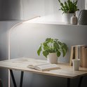Een gebogen, beige vloerlamp verlicht een houten bureau met een open boek en planten.