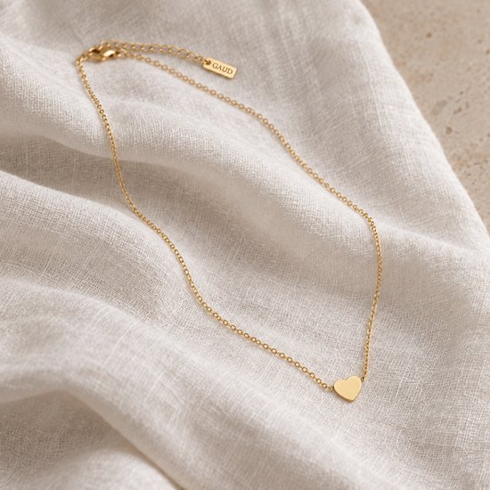 Gouden ketting met een klein, glanzend hartvormig hangertje op een dunne gouden schakelketting, liggend op witte stof.