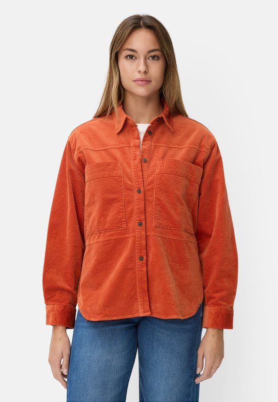Model draagt een oranje corduroy blouse met lange mouwen, overhemdkraag en twee borstzakken. Ze draagt een witte top en een spijkerbroek.