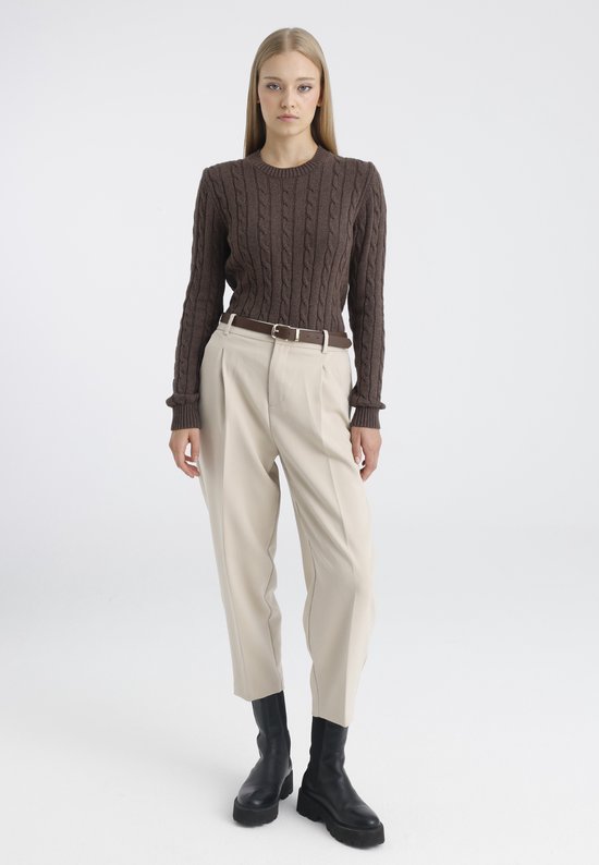 Model met bruine kabeltrui, beige broek, bruine riem en zwarte laarzen.