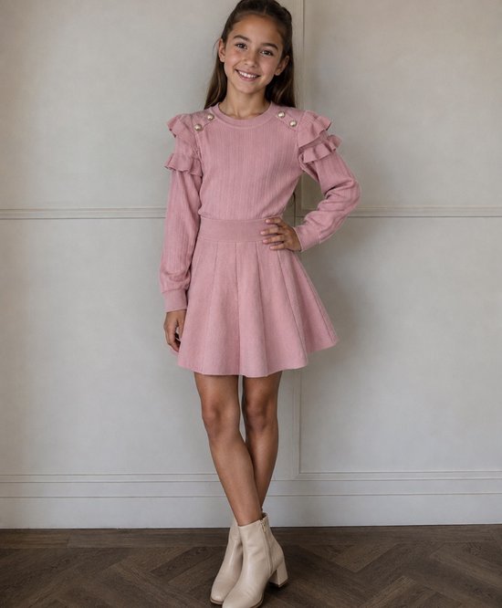 Meisje in een roze gebreide jurk met ruches en goudkleurige knoopjes op de schouders. De jurk heeft lange mouwen en een cirkelrok.