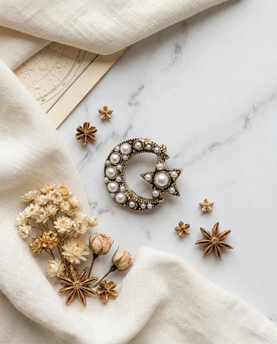 Gouden maan- en sterbroche ingelegd met parels, omringd door gedroogde bloemen op een marmeren ondergrond.