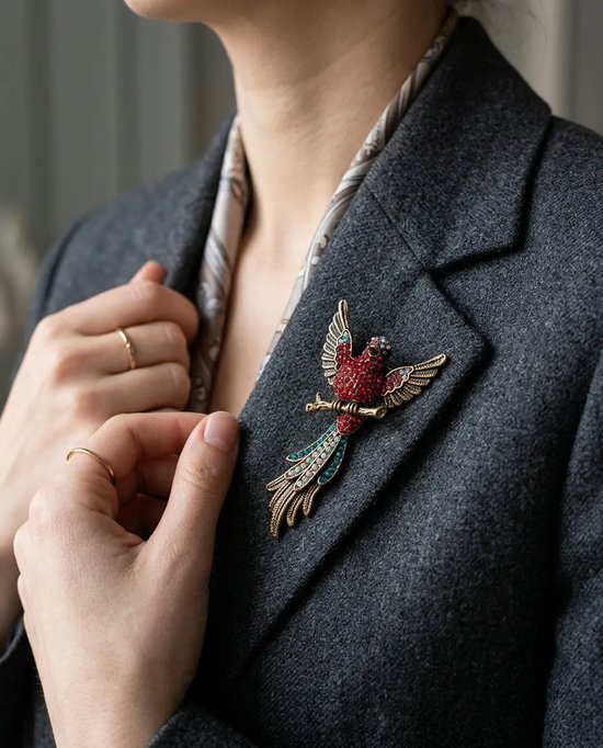 Gedragen antiekbronzen vogelbroche met fonkelende rode en blauwe stenen, gespeld op een grijze blazer.
