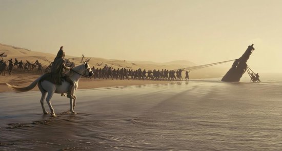 Ruiter op wit paard kijkt toe hoe een groep mannen een enorm object over een strand trekt tegen een zonsondergang.