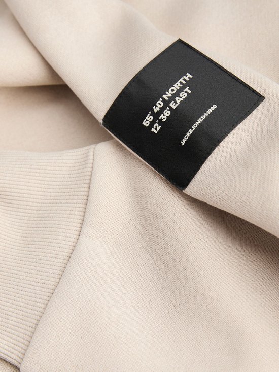 Detail van een zwart label met witte tekst op de mouw van een crème kleurige sweater. De tekst is '55° 40' NORTH 12° 36' EAST JACK&JONES©1990'.