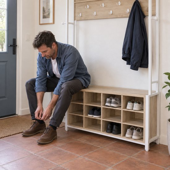 Man met bruine schoenen zit op schoenenrek, naast een kapstok met jas.