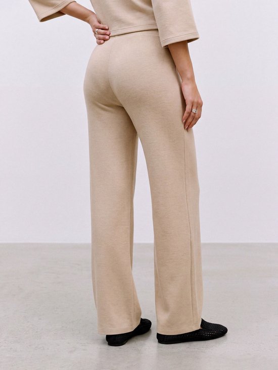 Een vrouw met zwangere buik, van achteren gezien, draagt een beige broek met wijde pijpen en een bijpassende top. Ze draagt zwarte schoenen.