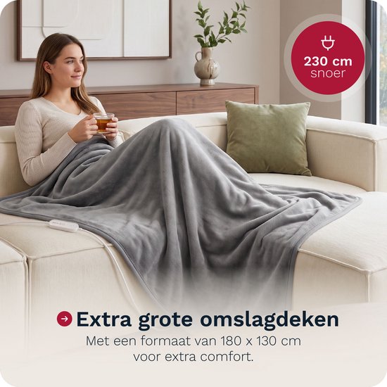 Vrouw op een bank met een grote grijze elektrische deken omgeslagen, met een kopje thee in haar handen.