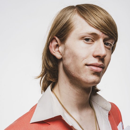 Jonge man met halflang blond haar, kijkt in de camera en draagt een koraalkleurig jasje met een gouden ketting.