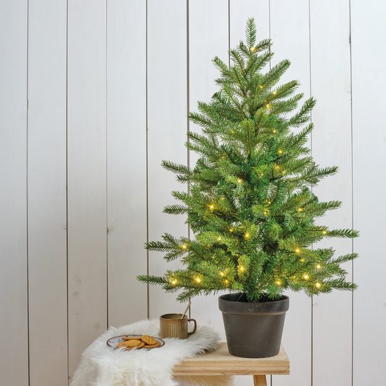 Kunstkerstboom in zwarte pot op een houten tafel naast een mok en koekjes, met warme verlichting.
