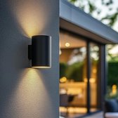Zwarte, cilindervormige wandlamp buiten die warm wit licht naar boven en beneden schijnt op een grijze muur.