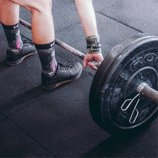 Persoon tilt een barbell op in de sportschool, dragend zwarte NObull-schoenen, sokken met camopatroon en polssteunen.