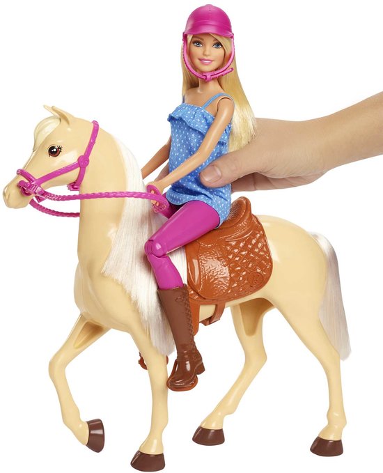 Een hand houdt een Barbie-pop vast die op een lichtbruin paard met roze helm zit.