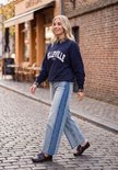 Model draagt een donkerblauwe trui met "BELLEVILLE PARIS"-tekst en een tweekleurige wijde spijkerbroek.