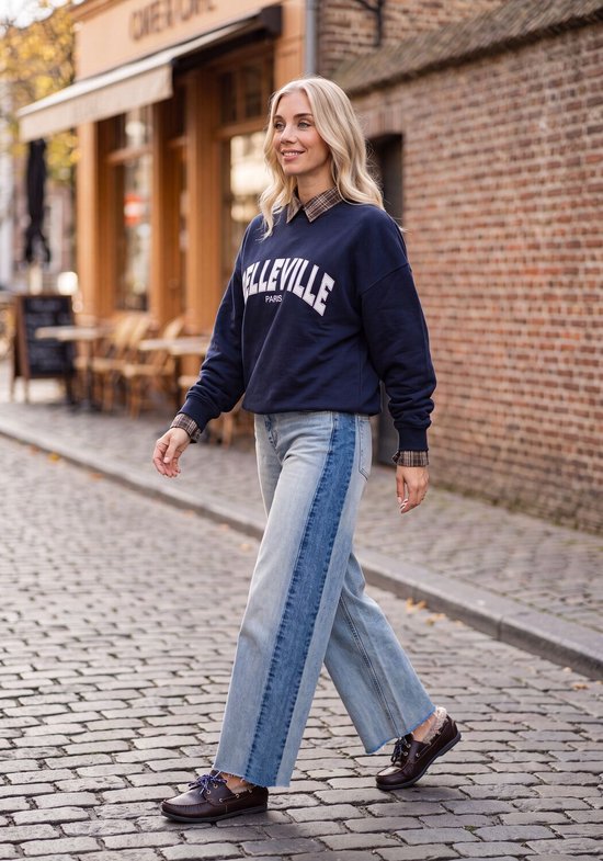 Model draagt een donkerblauwe trui met "BELLEVILLE PARIS"-tekst en een tweekleurige wijde spijkerbroek.