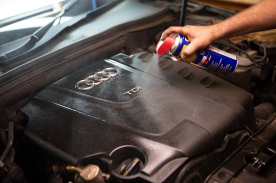 Handen gebruiken een WD-40 blik met rode spuitmond om een auto motor te behandelen.
