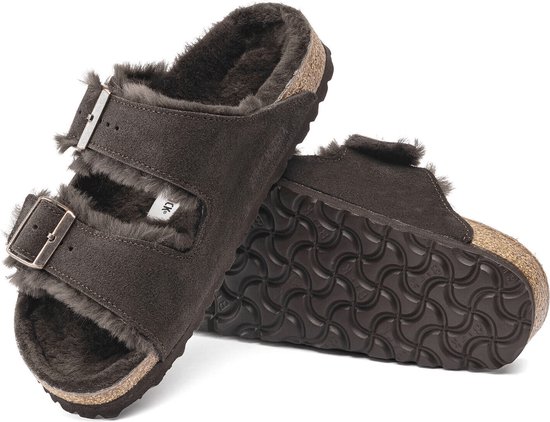 Een paar donkerbruine Birkenstock Arizona pantoffels, één van boven met gespen en lammenvacht, de andere van onder met profielzool.