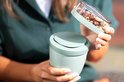 Een lichtgroene lunchpot met een transparant deksel vol met gemengde noten, die door een vrouw wordt vastgehouden.