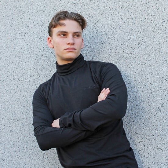 Jonge man in een zwarte coltrui met lange mouwen, leunend tegen een muur.