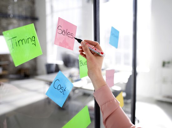 Hand met edding 3000 marker schrijft 'Sales' op roze post-it op glas.