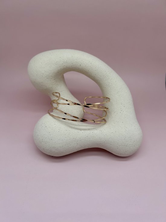 Gouden, gedraaide armband met golvend design op een abstracte, crèmekleurige standaard.