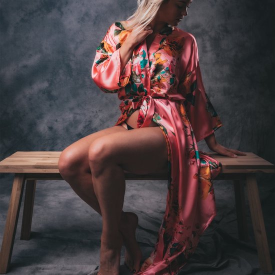Model zittend op houten bankje in koraalrode satijnen kimono met bloemenprint, hand op de nek.