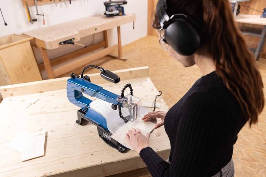 Vrouw met veiligheidsbril en gehoorbescherming gebruikt een blauwe Scheppach figuurzaagmachine.