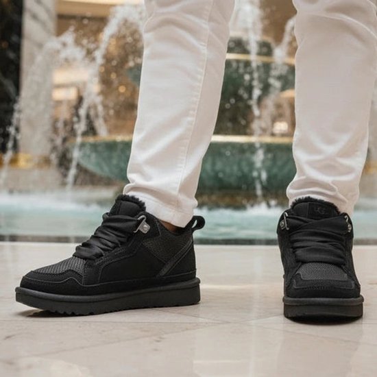 Man met witte broek draagt zwarte UGG sneakers met witte sokken, bij een fontein.