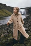 Model draagt een lange beige jas met reverskraag en opgestikte zakken, buiten in de natuur.