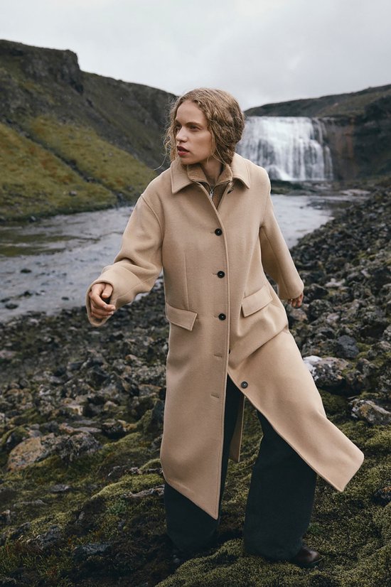 Model draagt een lange beige jas met reverskraag en opgestikte zakken, buiten in de natuur.