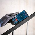 Grijze werkhandschoen die een Bosch laser afstandsmeter vasthoudt op een metalen leuning.