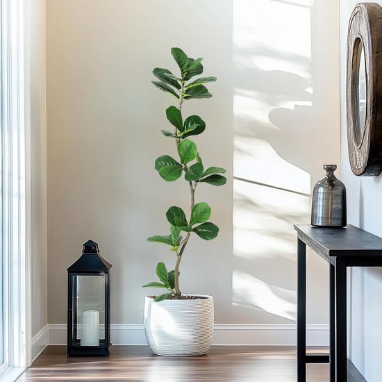 Een lange, groene kunstplant in een gestreepte witte pot, naast een zwarte lantaarn en een console.