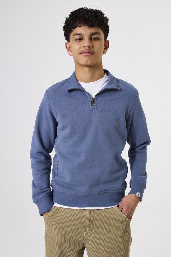 Jongen staat recht, draagt blauwe trui met rits, wit T-shirt en corduroy broek.
