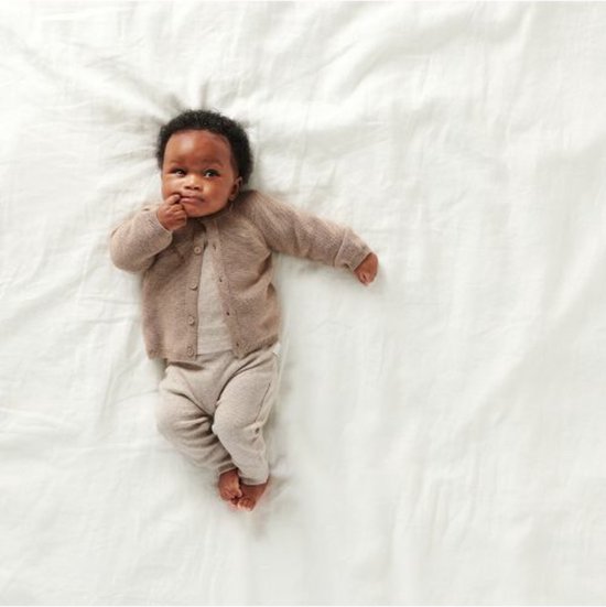 Baby ligt op een wit laken, een crème longsleeve shirt en een bruin vestje.