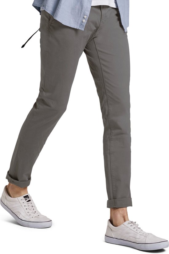 Man in grijze chino, opgerolde pijpen, witte sneakers en blauw hemd.
