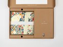 Une boîte en carton brun ouverte contenant une carte stylisée de bâtiments en bleu, orange et rouge sur fond crème.